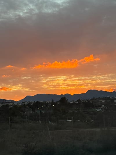 An evening scene reveals a striking sky ablaze with fiery orange and peach hues, contrasting dramatically with a darker, muted area to the left. Silhouetted mountains stretch along the horizon, partially illuminated by the warm light. Below the mountains, faint hints of a settlement with scattered lights are visible. A shadowy field occupies the lower portion of the image, creating a tranquil yet dramatic landscape.