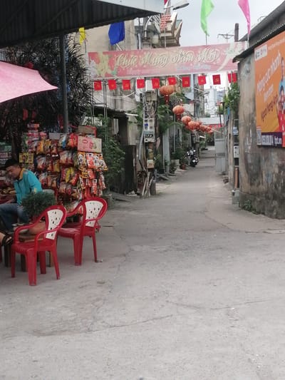 The image depicts a narrow street scene in Ha Tu Ward, Vietnam. A man in a blue shirt sits at a table next to a small storefront stacked with various packaged food items. Two red plastic chairs are positioned in front of the table. The street extends into the distance, lined with buildings and decorated with red flags bearing a communist symbol and a banner wishing a happy new year ("Chúc Mừng Năm Mới"). Red lanterns are strung above the street. Several parked motorbikes are visible further down the street. A billboard is attached to a wall on the right side of the street. The pavement is cracked and uneven.