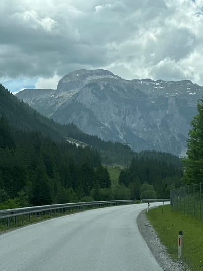 An overcast sky looms over a scenic mountain landscape in Flachauwinkl, Austria. A winding asphalt road curves through the frame, bordered by a metal guardrail on the left and a grassy embankment with a wire fence on the right. Dense evergreen forests cover the lower slopes of the mountains, giving way to rugged, rocky peaks in the distance. The mountains exhibit signs of erosion and have patches of snow on their upper reaches. A few power pylons are visible in the mid-ground, suggesting the presence of infrastructure. The scene is devoid of any people or animals.