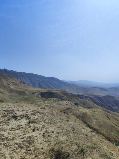 The image captures a wide, panoramic view of a hilly, arid landscape under a clear blue sky with sparse clouds. In the foreground, a dry, sandy slope is sparsely covered with dry grass and small green shoots. Beyond this, the terrain descends into rolling hills and valleys, characterized by dry, brownish-green vegetation and exposed earth. Several larger, darker mountains dominate the mid-ground and background, with rugged, rocky surfaces and steep inclines. Some areas of the hills show evidence of pathways or erosion channels. In the distance, further mountain ranges are visible, fading into a hazy blue, suggesting a vast, open geographical area. A small cluster of structures, possibly tents or temporary dwellings, and a few dark, indistinct shapes that could be animals, are visible in one of the lower valleys. The overall impression is of a remote, undeveloped natural environment. This scene is consistent with the terrain found in and around Sultanabad, Tajikistan, known for its mountainous and semi-arid regions.
