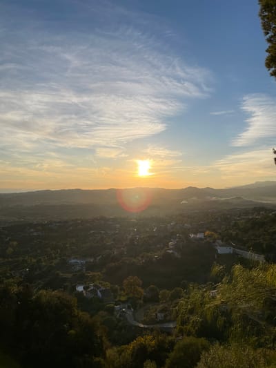 The photo captures a vibrant sunset viewed from a high vantage point. The sky is predominantly blue with wispy clouds, transitioning to a golden hue near the horizon. The sun shines brightly, creating a lens flare effect. Below, a lush, green landscape with scattered buildings and winding roads stretches into the distance, fading into shadows. The image is slightly rotated, offering a unique perspective of the scene.