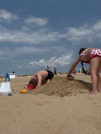 The image depicts a sandy beach scene, likely Vityazevo, Russia. In the foreground, a person wearing black swim trunks is partially buried in the sand, with their head facing down and a red bucket near their head. To their right, a yellow toy bucket is visible. In the mid-ground, another person, a woman, is wearing a strawberry-patterned bikini and is bending over. Her braid is visible, and she is holding a purple object. Further in the background, other people are present on the beach, including a person in a green swimsuit. The sky is blue with scattered white clouds, suggesting it is daytime and the weather is fair. The scene captures a moment of leisure and play on a sunny day at the beach. No visible text is present in the image.