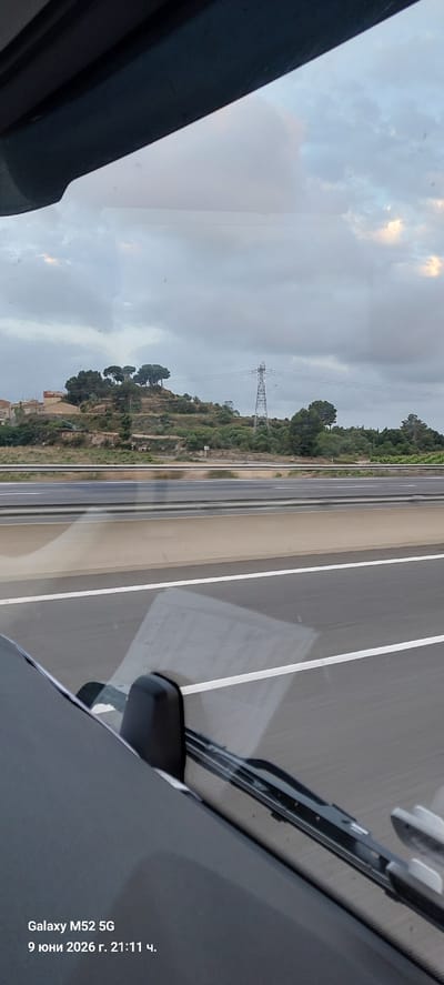The image depicts a view from inside a vehicle, likely a car, looking out through the windshield. The scene shows a highway with multiple lanes and a concrete barrier. On the right side of the highway, a hilly landscape is visible, covered with vegetation and trees. A large electrical pylon stands on one of the hills. A few buildings are also present on the hills. The sky is overcast with some patches of lighter clouds, suggesting either early morning or late afternoon. The bottom of the image shows the dashboard and windshield wipers of the vehicle, with text overlays indicating "Galaxy M52 5G" and "9 юни 2026 г. 21:11 ч." which translates to "June 9, 2026, 9:11 PM". Based on the context of the date and time, it is unlikely the image was taken in Banyeres del Penedès, Spain. The location indicated by the date and time is inconsistent with a typical daylight landscape view from a vehicle on a highway.