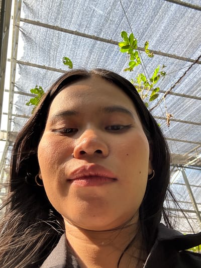 A low-angle, close-up, selfie-style photograph shows the face of a young person. The individual has dark hair, visible earrings, and makeup including eyeliner and lipstick. They are wearing a dark collared shirt. The background appears to be a greenhouse or protected outdoor area with a grey mesh covering overhead, through which sunlight filters and illuminates green leaves from a plant. The scene is located in Cileduk, Indonesia.