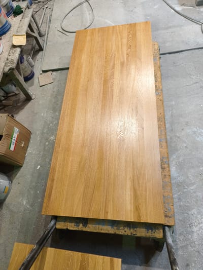 The scene depicts an interior workshop or industrial space in Negorci, North Macedonia. The primary subject is a large, rectangular wooden panel, likely a furniture component, exhibiting a light brown, golden hue with distinct vertical wood grain patterns. This panel rests horizontally on a dark, heavily stained wooden support structure, possibly a pair of sawhorses.

To the left of the central panel, a cluttered work area is visible. A smaller, stained wooden sawhorse supports multiple items, including several paint or solvent cans. One white can with blue lettering displays "SYNTHESIS SOLVENT BASED," and another partially visible label reads "MAXICOAT." A yellow rectangular piece of sandpaper, marked "P120" and "Smirdex," lies on the sawhorse. Below the sawhorse, a partially open cardboard box is positioned on the concrete floor, revealing a label with red, white, and green stripes and the text "UNIVERZAL NIF."

The floor throughout the visible area is concrete, gray, and shows signs of wear, splatters, and scattered debris. In the upper right quadrant of the image, a coiled black electrical cable or hose lies on the concrete surface. No individuals are present in the scene, and no specific actions or interactions are depicted. The environment suggests an active woodworking or finishing operation.