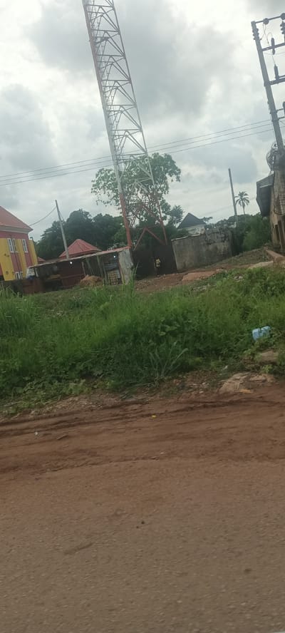 The scene presents an image captured in what appears to be a roadside setting within Umuoye, Nigeria. A large, lattice-structured telecommunications tower dominates the mid-ground. It's painted red and white at the base and extends upwards, its frame visible against an overcast sky.
Adjacent to the tower is a structure that seems to be a small shop or vendor's stall, constructed with corrugated metal sheets. The area around this stall is somewhat cluttered. A person is visible near the stall.
In the background, there are residential buildings of varying heights and colors. The architecture is simple, with some structures having corrugated iron roofs. The vegetation is lush and green, with trees and tall grasses populating the area.
To the right of the tower, power lines and utility poles stand alongside an older building that appears to be in a state of disrepair. The foreground is dominated by a dirt road, with visible tire tracks indicating vehicle movement. A blue plastic bottle is discarded on the side of the road. The image exhibits a slightly tilted perspective, which may have been shot from a moving vehicle.