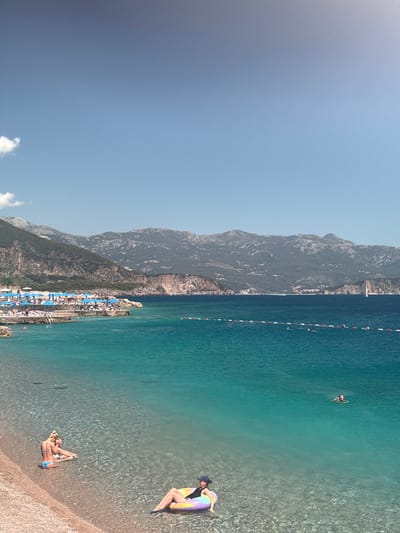 This detailed image captures a vibrant coastal scene in the city of Kubasi, Montenegro, under a clear, bright blue sky with a faint wispy cloud visible in the upper left. A rugged mountain range, primarily covered in dark green vegetation, forms the distant backdrop, with some lighter rock outcrops and scattered small buildings visible along its base and slopes.
The foreground features a light-colored pebbly beach that transitions into clear, shallow turquoise waters, revealing the seabed. On the left side of the pebbly beach, two individuals are seated close together in the shallow water, one with blonde hair and a visible light blue bikini bottom. On the right side of the foreground beach, another individual, wearing a dark top and a blue cap, is reclining on a multi-colored inflatable ring (purple, yellow, orange) in the shallow water.
Further out in the sea, the water deepens to a rich blue-green. A horizontal line of red and white spherical buoys marks a designated swimming area, extending from the middle-left towards the right side of the frame. Several people are swimming within and beyond this marked area, including one individual visible swimming in the mid-ground, right of center.
To the left, a developed beach area or pier is visible, densely populated with numerous blue beach umbrellas and sunbeds, accommodating many patrons. In the far distance on the right, a white sailboat with a single mast is discernible against the water and mountains. The sun appears to be positioned from the upper right, casting a bright illumination across the scene.
