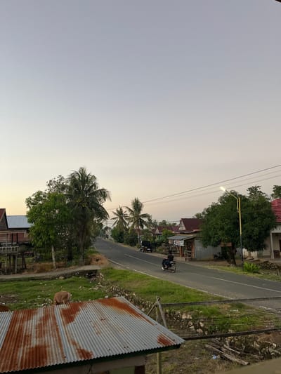 The media file depicts an outdoor scene in Turumame, Indonesia, likely during the late afternoon or early evening. The sky is a pale, hazy yellow-orange, indicating either dawn or dusk. A paved road runs diagonally through the scene, bordered by lush green grass and vegetation.

Several houses with red roofs are visible along the road, suggesting a residential or semi-rural area. Tall trees, including palm trees, are scattered throughout the landscape. A street lamp with a lit bulb stands near the road, casting a glow.

In the foreground, on the right side of the image, is a corrugated metal roof, showing signs of rust. A light brown animal, possibly a dog or a calf, is lying on the grass next to the roof, facing away from the camera.

Further down the road, a person is riding a motorcycle. The person is wearing dark clothing. Other motorcycles are parked near some of the houses.

The overall impression is one of a peaceful, everyday scene in a tropical or subtropical location. The lighting suggests a gentle end to the day. There is no visible text in the image.