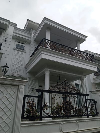 An ornate two-story building features white exterior walls, prominent columns, and detailed decorative accents. A wrought-iron fence with elaborate gold-toned embellishments surrounds the property. Balconies with equally ornate railings extend from the upper floors. The architecture displays a classical style with a touch of modern design. The sky is overcast.