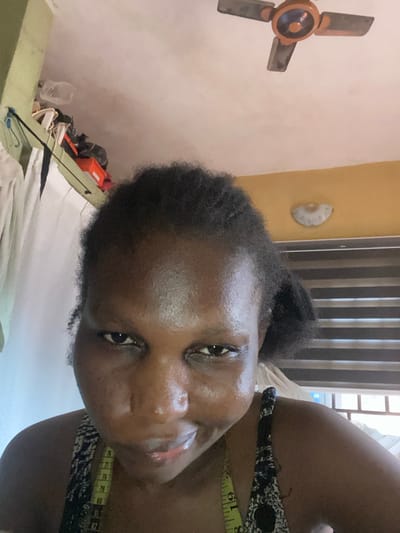 The image captures a close-up portrait of a person, appearing to be a Black woman, in an indoor setting in Oba-Aboji, Nigeria. Her head is tilted, and her face, with noticeable sheen, occupies a significant portion of the frame. She has dark, curly hair pulled back. She wears a dark-patterned top or dress, and a yellow measuring tape is draped over her right shoulder/neck, showing the number "19".
The background indicates a domestic environment. To the viewer's left, a light-colored wall features a brown and orange three-bladed ceiling fan. Above and behind the person's head, a light-colored, shell-like decorative object is affixed to a yellow wall section. A window with vertical blinds is visible further back, allowing natural light into the room, suggesting it is daytime. In the lower left, there's a cluttered area on a surface, including an orange box and various items, partially covered by a white sheet or fabric. The person's expression appears relaxed or neutral, capturing a casual moment, possibly a selfie.