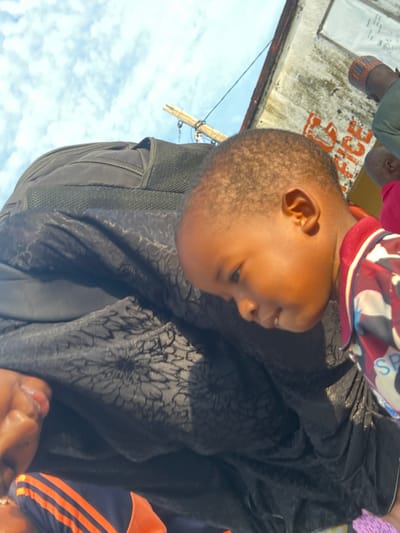 The image presents an outdoor scene in Gbaza, Nigeria, captured from a low, tilted perspective. A young child, approximately a toddler, with short dark hair and light brown skin, is prominently featured on the right, looking downwards and to the left with a slight smile. The child is wearing a collared shirt with red, white, and blue geometric patterns, and the partial letters "SP" are visible on the fabric. The child appears to be held or carried by an adult, whose upper back and left shoulder are visible, covered by a black garment with a subtle, dark textured pattern. A black backpack with a zippered compartment and mesh shoulder straps is visible on the adult's back. Below the black garment, an article of clothing with distinct orange and blue stripes is discernible. In the background, an weathered, light-colored wall or building surface is visible, featuring red painted letters that partially spell what appears to be "ICE" or a similar word, along with a white rectangular sign affixed to it. Portions of two other individuals are visible in the upper right background: one wearing a green garment and a patterned cap, and another person dressed in pink. The upper left portion of the frame shows a blue sky with scattered white, wispy clouds and a segment of a wooden pole or structure extending from the left.