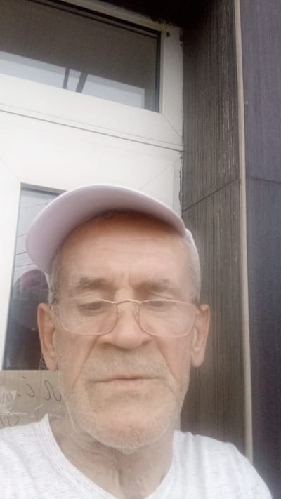 The image is a close-up selfie of an elderly man wearing a white cap and glasses. He is outdoors, with a white-framed window visible behind him on the left and a textured, dark grey wall on the right. The lighting suggests it is daytime, possibly overcast. The man has fair skin, graying hair, and a light stubble. He appears to be looking down and slightly to the side, with a neutral expression. His clothing is a white, textured t-shirt. There are some faint, dark scribbles on a surface visible below and to the left of the man, but no clear text is discernible. The overall impression is a casual, everyday portrait taken in an outdoor setting.