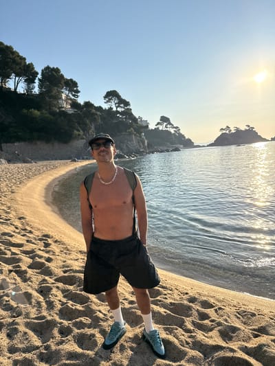 A man stands on a sandy beach in Platja d'Aro, Spain. He is shirtless, wearing black shorts, white socks, and blue athletic shoes. He has a silver chain around his neck and is wearing a baseball cap and sunglasses. A backpack is slung over his shoulders. The beach has footprints in the sand. In the background, the ocean is calm with sunlight reflecting on the water. There are trees and buildings on a hill overlooking the beach. The sky is clear and blue with the sun setting, casting a warm glow on the scene.