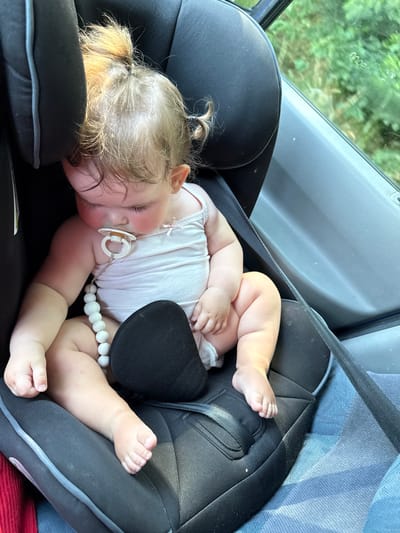 The image shows a baby sleeping peacefully in a car seat. The baby is lying on their side, with their head resting on a black cushion. They are wearing a light pink onesie and have a pacifier in their mouth, attached to a white beaded clip. The baby's chubby arms and legs are visible.
The car seat is black with grey trim and a black harness strap is visible across the baby's body. The background shows a car interior, with a grey door panel and a hint of blue fabric. Through the window, blurred green foliage is visible, suggesting the car is in a natural setting. The lighting indicates it is daytime, likely with sunlight filtering in.
The scene conveys a sense of calm and tranquility, with the sleeping baby appearing comfortable and secure. The image was taken in Volpertshausen, Germany. There is no visible text in the image.
