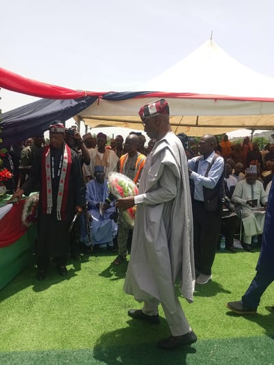 Captured in Dilpurtu, Nigeria, the image shows an outdoor gathering, likely a funeral or memorial event. A man in a gray robe and cap carries a wreath, walking on artificial turf. Behind him, a man in a black suit and patterned sash stands near a draped coffin, possibly containing the remains of the deceased. A photographer is capturing the scene. A seated man in a blue robe holds an automatic rifle. Other attendees are visible in the background under a tent.