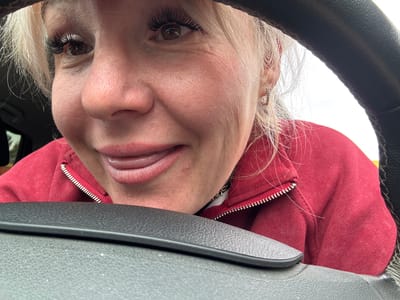 The image shows a close-up, low-angle, selfie-style shot of a fair-skinned Caucasian woman in her late 30s to early 40s, with blonde hair and full eyelashes, leaning her head on a steering wheel. She is wearing a red hooded sweatshirt with a zippered front. Her mouth is slightly open, and her tongue is visible, touching her upper lip. She is wearing small hoop earrings. The background appears to be a car interior, with the steering wheel in the foreground. The lighting is bright, suggesting it's daytime. The image is taken from inside the car, looking outwards. There are no discernible landmarks or environmental features that would indicate the specific location of Chastyye, Russia.