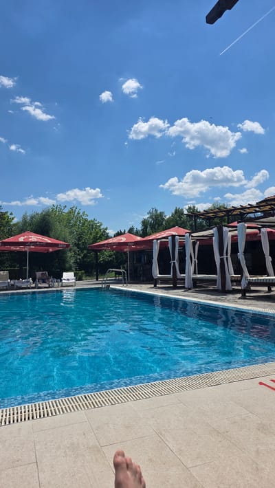 The image shows a swimming pool area on a sunny day. In the foreground, a person's bare foot is visible on the tiled edge of the pool. The pool itself is filled with clear blue water. To the left of the pool, several lounge chairs are set up, with one person relaxing on a chair. Several large red umbrellas, branded with "1859", provide shade over the lounge area. Further to the right, there are several cabana-style seating areas with white curtains. In the background, lush green trees form a dense backdrop under a bright blue sky dotted with fluffy white clouds. A contrail from an aircraft is visible in the upper right portion of the sky. The scene depicts a resort or recreational area, possibly at the hotel in Simeonovo, Bulgaria, given the context provided.