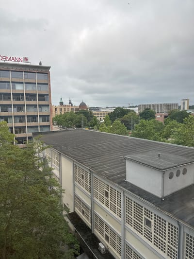 The image captures an urban cityscape under an overcast sky, likely in Chemnitz, Germany, given the visible architectural styles and signage. In the foreground, the flat, dark rooftop of a building with a prominent ventilation unit is visible. Adjacent to this, the side of another building with a white facade and a grid-like pattern of windows is prominent. Further into the background, a taller, multi-story building with a beige facade and rows of windows stands on the left. The roof of this building displays red lettering that reads "ÖORMANN" and a logo. Two figures appear to be standing on the roof of this building, near the lettering. Beyond these buildings, a cityscape unfolds with various structures, including buildings with domed roofs, trees, and what appears to be a crane in the distance. The overall atmosphere is muted due to the cloudy weather.