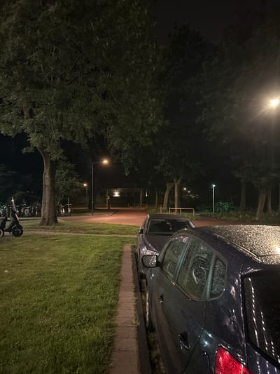 The image depicts a nighttime street scene in Leiden, Netherlands. Two dark-colored cars are parked side-by-side on a paved area, with the car in the foreground showing numerous water droplets on its surface, suggesting recent rain. Adjacent to the parking area is a strip of grass, and beyond that, more grass and trees line the street. In the background, a row of parked bicycles and scooters is visible under the illumination of streetlights. Further in the background, a building with illuminated windows can be seen, and more trees and streetlights are scattered throughout the scene. The overall atmosphere is dark, with the primary light sources being streetlamps and the building's interior lights, creating a slightly hazy and reflective environment due to the wet conditions.