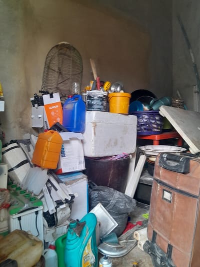 In Gauraka, Nigeria, a cluttered room contains various items piled high. A large, dark brown suitcase sits on the right side of the frame. To its left, a dark grey trash bag is partially visible. Several white and light grey boxes are stacked together, with some appearing to be storage containers.
On a lower level, a light brown plastic jerrycan is present, as is a smaller white plastic bottle with a red cap. A bright blue plastic container with a black handle is positioned on top of other items. An orange plastic jerrycan is also stacked amongst the boxes.
In the background, a large, dark wire cage or fan guard is positioned against a textured, beige wall. Various tools, including what appear to be knives and a hammer, are visible near the top of the frame. Other assorted objects, including buckets, baskets, and what looks like a white board or panel, are scattered throughout the scene, suggesting a storage or workshop area. The lighting is somewhat dim, and the overall impression is one of disarray and accumulation of goods.
