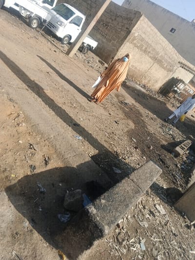 This image depicts a dusty, unpaved street or open area in Gobirawa, Nigeria, under bright daylight with strong shadows indicating an angled sun.

In the midground, slightly left of center, a person stands facing towards the right foreground. This individual wears a light tan full-body garment and a light blue surgical face mask, covering their head and face except for the eyes. They hold a white, rectangular object in their left hand, resting it against their body. Their feet are visible in open-toed sandals. Behind this person and to their left, a utility pole casts a long shadow diagonally across the ground towards the right foreground. The pole has red markings. Further to the right and slightly behind, a second person is partially visible, dressed in a long white garment, facing away from the camera. A yellow object is visible on the ground near their feet.

The ground is predominantly packed dirt and dust, uneven, and littered with various pieces of refuse including plastic bags, paper, and small debris. In the immediate foreground, a broken concrete structure, possibly part of a curb or drain, contains more debris.

In the background, on the left, several white compact pickup trucks are parked on the unpaved surface. Beyond them and across the entire background are buildings constructed of unplastered cinder blocks. One prominent wall displays graffiti, including "AN AKI" and the sequence "5303222". To the far right background, near another block building, rudimentary wooden shelving or structures are visible. The sky is clear and bright at the top of the frame.