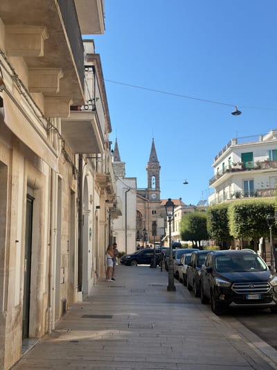 The image depicts a street scene in Alberobello, Italy, on a clear, sunny day. On the left, a light-colored building with classical architectural elements, including columns and balconies, lines the street. A woman in a white top and shorts stands on the sidewalk, looking towards a store entrance. Across the street, a row of parked cars, including a black SUV with an Italian license plate FP-766HT, are visible. Further down the street, a white building with balconies adorned with potted plants stands out. In the background, a tall bell tower with a clock face rises above the other structures, indicative of a church or cathedral. The sky is a bright, cloudless blue. The ground is paved with cobblestones, and lampposts are spaced along the sidewalk. Lush green trees are planted along the street, providing shade.
