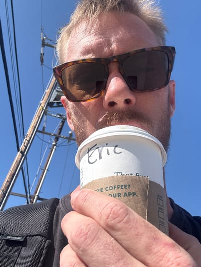 A selfie capturing a man with sunglasses drinking from a white coffee cup labeled "Eric." The cup features a brown corrugated sleeve. The photo is taken from a low angle, looking up at the man against a bright blue sky. Power lines and wooden poles are visible in the background. The man's hand holding the cup and part of a black bag or clothing are also in view.