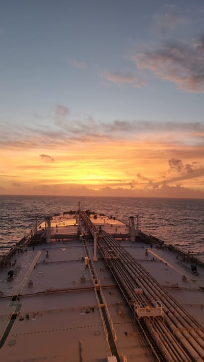 The image shows a wide shot of a large cargo ship at sea during sunset. The sky is filled with vibrant colors of orange, yellow, and pink from the setting sun, with streaks of clouds creating a dramatic effect. The sea is calm and reflects the colors of the sky. The deck of the ship is visible, with numerous pipes running along its length, typical of an oil tanker or chemical carrier. There are no people visible on the deck. The overall impression is one of peacefulness and solitude, with the vastness of the ocean and the beauty of the sunset. The time of day is clearly sunset, and the weather appears clear with some scattered clouds. The location is at sea, on a cargo ship. There is no visible text in the image.