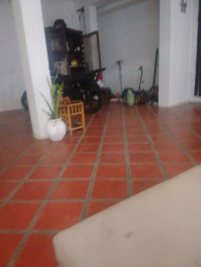 The image depicts an indoor scene with a tiled orange floor. A white pillar is on the left side. In the background, there is a dark wooden cabinet with glass doors. To the right of the cabinet, a motorcycle is parked. A small, wooden stool is next to a white, round vase containing a green plant. Further back, several objects are visible, including what appear to be canes or walking sticks and some green material on the floor. The lighting in the scene is dim. This image is located in Atamo Sur, Venezuela.