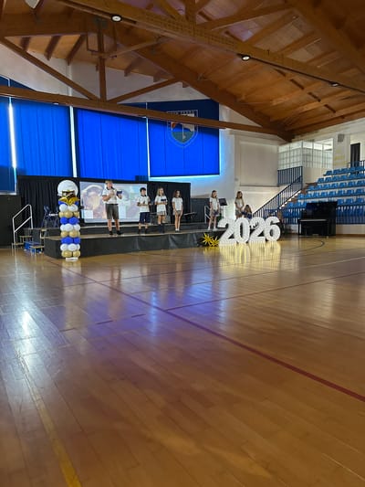 This photograph captures a graduation or recognition ceremony taking place in what appears to be a school gymnasium. The scene is dominated by a polished wooden floor, with various lines indicating court markings. A raised stage is set up on the left side of the image, draped with black fabric. On the stage, a group of students, dressed in school uniforms, are gathered. Some are standing and holding what look like certificates or awards, while others are seated. A large screen is visible behind them, displaying an image, possibly related to the school or the event.

In front of the stage, large, illuminated numbers spelling out "2026" are a prominent decorative element, suggesting this is an event for the graduating class of that year. Beside the numbers, a cluster of gold and blue balloons adds to the festive atmosphere. A microphone stand and what looks like a keyboard or piano are also present on the stage area.

In the background, blue bleacher seating is visible, indicating a space designed for an audience. A grand piano is positioned near the bleachers. Further back, a staircase leads up to an elevated area. The ceiling features exposed wooden beams and beams, suggesting an older, perhaps traditional architectural style for the gymnasium.

The lighting suggests an indoor event, possibly in the evening or a dimly lit space. The overall mood appears celebratory and formal. On the blue backdrop behind the stage, a crest or logo is visible, and below it, text that partially reads "SAN PEDRO". The word "WEST HOUSE" is visible on a sign near the top of the bleachers.