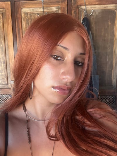A person with auburn hair and brown eyes poses for a close-up shot. They have a light complexion with a subtle lip gloss. A silver hoop earring is visible, along with a silver choker and a necklace with small beads. The background features a wooden cabinet with decorative carved panels and a metal garment hanger.