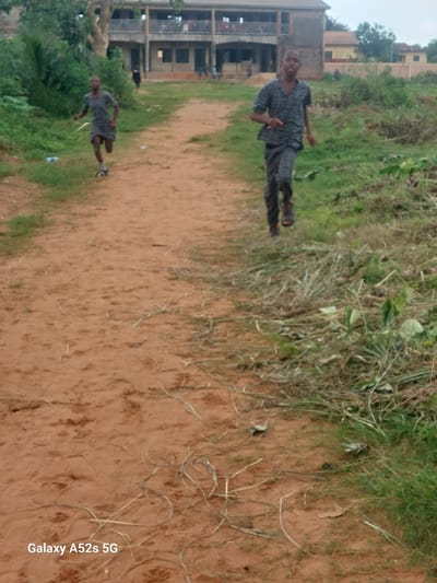 Two young boys are running on a dirt path in Umudioka, Nigeria. The boy in the foreground, wearing a checkered shirt and pants, is running towards the camera. He is wearing sandals. The second boy, in the background and to the left, is also running away from the camera. He is wearing a short-sleeved shirt and shorts. In the background, there is a two-story building with balconies and windows. The area around the path is covered in grass and dirt, with some vegetation visible. The sky is overcast.