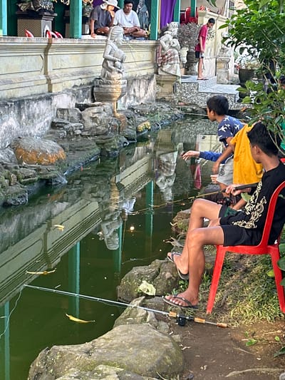 In Bangli, Indonesia, two boys are fishing in a pond. The boy on the left is wearing a blue shirt with white bird patterns and grey shorts. He is leaning over the pond, looking into the water with a fishing rod in his hand. The boy on the right is wearing a black long-sleeved shirt with gold designs and black shorts. He is sitting on a red plastic chair, also holding a fishing rod and looking towards the pond.
The pond has green, murky water, with rocks and some fallen leaves on its surface. Several fishing rods are visible, cast into the water. The background shows a building with a stone facade and decorative statues, suggesting a temple or a traditional complex. Trees and lush greenery are also present, indicating an outdoor setting. The lighting suggests it is daytime, possibly late afternoon, with sunlight casting reflections on the water.