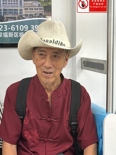 An elderly Asian man wearing a straw cowboy hat that says "ronaldino" across the front is sitting on what appears to be public transportation, possibly a train or bus. He is wearing a maroon, mandarin-collared shirt with dragon embroidery and traditional knot closures. A black backpack is visible on his shoulders. The background shows a wall with a large advertisement featuring a building and Chinese text, as well as a red emergency sign with a prohibition symbol and text in Chinese and English. The man is looking to his right with a slight smile. The environment appears to be indoors, and given the text on the advertisement and the emergency sign, it is likely a public space in China. The text "Tiaodeng, China" cannot be confirmed from the image alone.