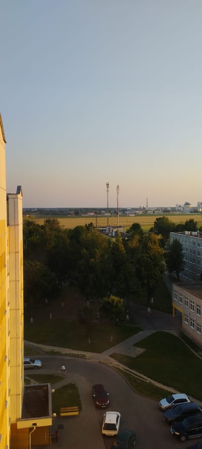 The image depicts a view from a residential building in Skidzieĺ, Belarus, showcasing a street scene with parked cars and a few individuals. The time of day appears to be late afternoon or early evening, indicated by the warm, soft lighting and long shadows.
In the foreground, the side of a yellow building is visible, suggesting it is a multi-story apartment complex. A paved courtyard area is visible below, with several cars parked. A silver sedan is parked on the left. Further into the courtyard, a dark red SUV is parked, and in front of it, a white hatchback. To the right, a dark blue sedan and a black SUV are parked. Two small figures, possibly children, are standing near the silver car, one of them seemingly on a scooter.
A yellow park bench is situated on the paved area, adjacent to a building entrance. The courtyard leads to a winding road.
In the mid-ground, a dense line of green trees separates the residential area from a field. Beyond the trees, two tall communication towers with red and white markings stand prominently against the sky. In the distance, industrial buildings and structures, including a tall red and white chimney, are visible, suggesting an industrial zone or the outskirts of the city. The sky is clear and a pale blue, with a gradient towards a warmer hue near the horizon. The overall scene conveys a sense of a quiet urban residential environment on a clear day.