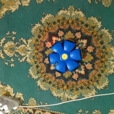 An overhead, medium shot shows a blue, petal-shaped toy with a yellow center resting on a teal carpet with an ornate, multi-colored pattern. The toy is positioned in the center of the image, with its petals spread outwards. A thin white cord extends from the bottom left corner of the frame towards the toy, and a grey and white object, possibly a part of a camera or lighting equipment, is visible in the bottom left corner, partially obscured. The carpet's pattern features floral motifs in shades of yellow, orange, and cream against the teal background. The lighting appears to be artificial, casting a soft glow on the scene. This image was taken in Almaluu, Kyrgyzstan.