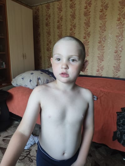 A young boy with a shaved head and light skin stands in what appears to be a bedroom. He is wearing dark blue briefs. His chest is bare. His left arm shows a noticeable tan line. Behind him, a bed with an orange cover is visible, along with a pillow with a blue floral pattern. The wall behind the bed is covered in a cream-colored wallpaper with a repeating floral pattern in shades of brown and red. To the left of the frame, a portion of a wooden cabinet or closet is visible. The lighting suggests it is daytime. The boy's expression is neutral, with his mouth slightly open. There is no visible text in the image. The location is Askiz, Russia.