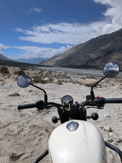 A first-person perspective shot of a white motorcycle is positioned in a rocky, sandy terrain, overlooking a vast, arid valley. The motorcycle's handlebars, mirrors, and part of its fuel tank are visible in the foreground. The left handlebar has a yellow carabiner attached to it. The background reveals a wide riverbed, with sparse vegetation and rocky slopes leading up to imposing, rugged mountains under a bright blue sky dotted with white clouds. The environment appears to be a remote, mountainous landscape, possibly in the region of Sasoma, India, given the user's prompt. No people are present in the image. The scene is static, with no apparent actions taking place. The lighting suggests it is daytime.