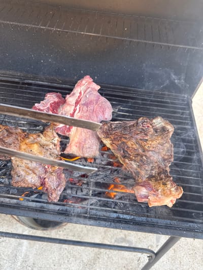 In Dunya, Bulgaria, on a sunny day, three cuts of raw beef are being grilled over hot coals on a black metal barbecue. Two pieces of meat, one larger and thinner, the other thicker and marbled, are positioned on the right side of the grill, with the larger piece showing a well-seared crust. To the left, two more pieces of raw beef are being held by silver metal tongs, one of which is also positioned over the coals. Wisps of smoke rise from the grill, indicating the cooking process is underway. The barbecue is situated outdoors, with a hint of a light-colored concrete surface visible beneath it.