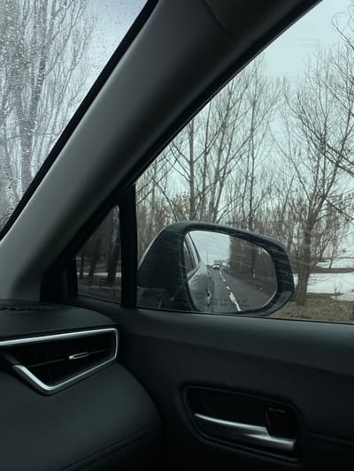 The image presents a view from the interior of a vehicle, likely in or near Solak, Armenia. The perspective is directed towards the front passenger side, encompassing the vehicle's dark dashboard with a chrome-trimmed air vent, a dark door panel with a chrome handle, and the A-pillar. The exterior window is visibly speckled with raindrops.

Through the window and reflected in the side-view mirror, a winter or early spring landscape is visible under an overcast, light grey sky. Numerous bare deciduous trees, stripped of leaves, line the roadside. The side mirror clearly reflects a two-lane paved road with white lane markings, where a grey sedan is observed directly behind the camera's vehicle, and a truck is visible further down the road in the distance. The mirror's surface displays the warning "OBJECTS IN MIRROR ARE CLOSER THAN THEY APPEAR." To the right of the road, beyond the trees, the ground shows patches of snow or ice, bordering a body of water. No individuals are visible in the scene.