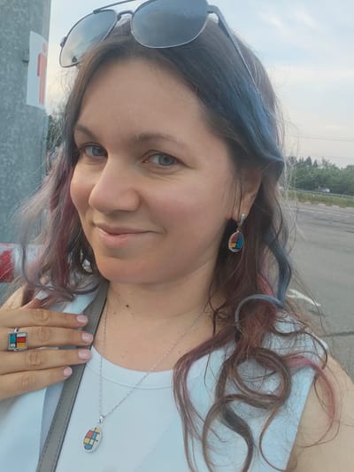 A medium close-up shot features a woman with long, wavy brown hair with streaks of blue and pink, wearing sunglasses perched on her head. She is smiling and looking towards the camera. She wears a white tank top and a necklace with a pendant. On her left ring finger, she wears a ring with a colorful geometric design, matching her earrings and necklace. The background appears to be outdoors with trees and a road visible, suggesting a location in the city of Sholokhovo, Russia. A sign with a red silhouette of a person is visible on a metal pole to the left of the woman.