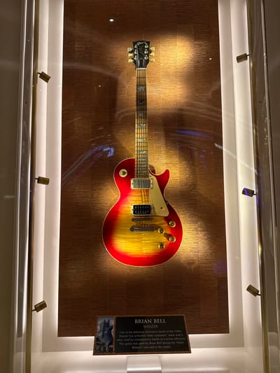 The image captures Brian Bell of Weezer's guitar displayed in a case. The guitar, a Gibson Les Paul with a red and yellow sunburst finish, is mounted against a textured, metallic gold background. Above and below the guitar are small track lights illuminating the display. To the right of the guitar is a plaque with information about Brian Bell and Weezer. The text on the plaque mentions Weezer's influence as a 1990s band and states that the guitar was used during the "Make Believe" tour and in the studio. The photograph was taken at the city of Hollywood Seminole Reservation, United States of America.
