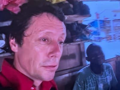 The image depicts two male individuals in an indoor setting within the city of Colombes, France. In the foreground, positioned to the left, is a light-skinned male with dark, wavy hair, visible wrinkles on his forehead and around his eyes. He is wearing a red collared shirt and exhibits a neutral, slightly contemplative expression, with his gaze directed downwards and to the right. Behind him and to the right, partially obscured, is a second male, appearing younger, with dark skin and short, dark hair. He is dressed in a light-colored, patterned t-shirt, and his face is partially shadowed. The background features horizontal wooden planks, suggesting a wall or structure, with an indistinct, blurry area above that includes shades of blue and white, possibly distorted reflections or out-of-focus elements. No direct interaction or specific action is discernible between the two individuals. The image quality suggests a photograph of a digital screen.