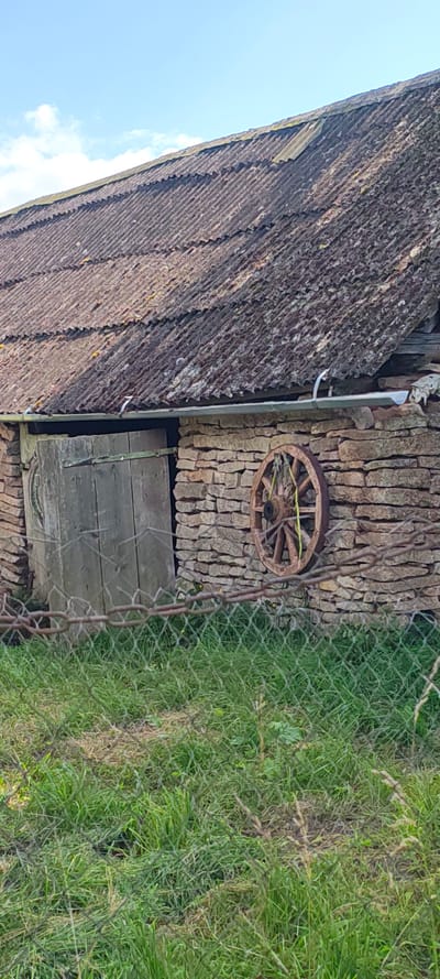 The image depicts the exterior of a rustic stone building, likely a barn or shed, located in the village of Malaya Listovka, Russia. The building's roof is made of corrugated material, weathered and covered in moss. A wooden door, appearing aged and weathered, is visible on the left side of the structure. To the right of the door, mounted on the stone wall, is a wooden wagon wheel, adorned with green straps. The building is situated behind a wire fence, with green grass growing in front of it. The sky above is blue with scattered white clouds.