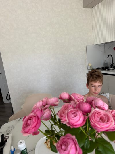 In a brightly lit, modern kitchen or dining area, a young male with short, light brown hair is seated at a table, looking downwards. He is shirtless, with a light-colored plaid garment draped over his shoulders. In the foreground, a bouquet of vibrant pink roses is arranged in a vase, partially obscuring the table and the lower portion of the individual. The table itself is round with a marble-patterned surface. On the table, several items are visible: a white box, a watch, a bottle with a label, and a plate with a folded pastry, possibly a crepe or pancake. The background features a textured light-colored wall and a section of a white kitchen counter with a black faucet and a bottle of soap dispenser. The overall setting suggests a domestic interior.