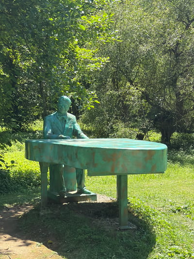 This media file shows a statue of a man sitting at a table in an outdoor setting. The statue is made of a material that appears to be aged bronze, with a greenish patina and some reddish-brown accents. The man is depicted wearing a suit and appears to be in a pensive pose, with his hand resting on the table. The table is large and rectangular, also with a greenish patina.
The statue is situated in a lush green environment, with trees and grass surrounding it. The sunlight suggests it is daytime, likely late morning or early afternoon, casting shadows on the ground. The overall impression is one of a serene and artistic display within a natural landscape. The location is identified as Shekvetili, Georgia. There is no visible text in the image.