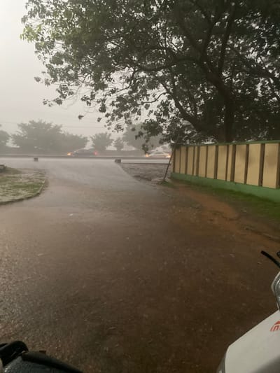 The image shows a street scene in Jama A, Nigeria, during a heavy rainstorm. The road is wet and shiny from the rain, and visibility is reduced due to the heavy downpour and fog. There are trees along the left side of the street. A vehicle with headlights on is visible further down the street. The presence of a building on the edge of the frame suggests the setting is an urban or suburban area. The image has been rotated, with the original horizontal view now oriented vertically. A portion of a motor vehicle is visible in the upper right of the frame.
