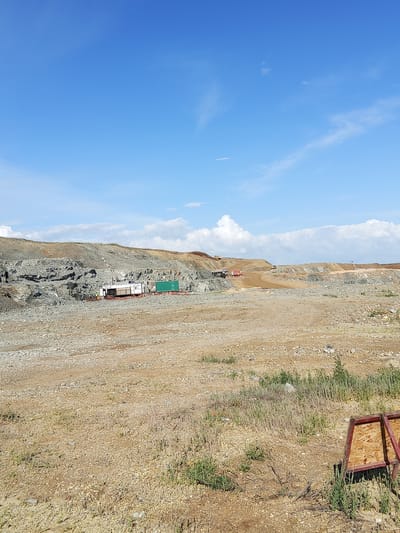 This media file depicts an outdoor construction site under a bright blue sky with scattered white clouds. The scene is a quarry or excavation site with rugged, rocky terrain and large piles of excavated material. In the mid-ground, there are several pieces of heavy machinery, including what appears to be a drilling rig, a white truck with a trailer, and a green shipping container. A small red and white vehicle is also visible. A person in a yellow vest is standing near the drilling rig. The ground is mostly dry and dusty, with patches of sparse vegetation. In the background, the terrain rises, revealing layered rock formations and further areas of excavation. Several yellow construction vehicles, likely excavators or bulldozers, are working in the distance, shaping the landscape. The overall impression is one of active construction or resource extraction. The lighting suggests it is daytime, likely late morning or early afternoon, given the clear sky and shadows. There is no visible text in the image.