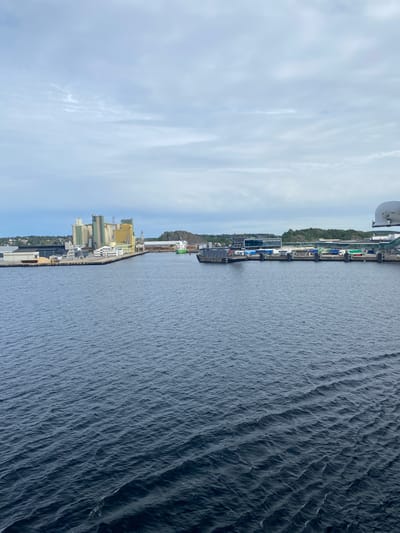 The image shows a port in Larvik, Norway, with a body of water in the foreground and industrial buildings along the coastline in the background. The water is dark blue and appears to have small waves. The buildings are a mix of modern and industrial structures, with some appearing to be grain silos or storage facilities. There are also some cranes visible, suggesting ongoing activity at the port. The sky is overcast with some clouds. The time of day appears to be daytime, given the lighting. The overall scene is industrial and functional, typical of a working port. There is no visible text in the image.