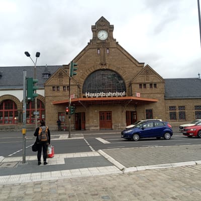 The image depicts the exterior of the Hauptbahnhof (main train station) in Eisenach, Germany. The architecture of the building is characterized by rough-hewn stone blocks and a large, arched stained-glass window above the entrance. A clock is prominently displayed on the upper facade of the building. In front of the station, a traffic light is visible, currently showing a red signal. A woman with a backpack and shopping bags stands on the sidewalk, facing away from the camera, waiting to cross the street. Several cars are stopped at the traffic light, including a blue hatchback, a white sedan, and a red compact car. The sky is overcast. The street in front of the station is paved with cobblestones, and there is a pedestrian crossing marked on the road. The overall scene suggests a typical urban environment on a cloudy day.